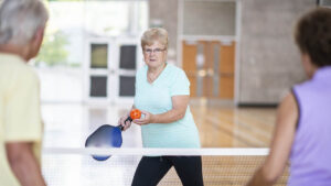 woman serves the ball playing Pickleball. two other people wait to receive the serve